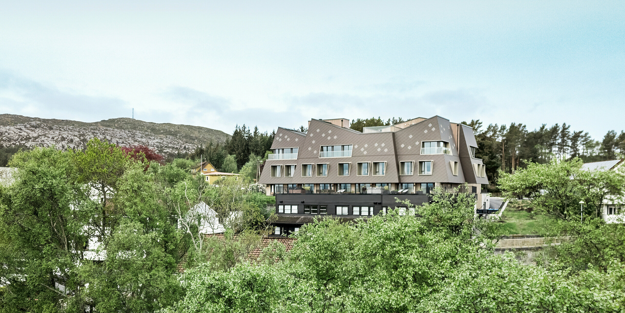 Blick aus der Ferne auf das Boutiquehotel Beckerwyc House in Bekkjarvik, Norwegen, eingebettet in eine grüne, hügelige Landschaft. Das futuristisch anmutende Gebäude ist mit PREFA Wandrauten 44 × 44 in Nussbraun verkleidet, die sich harmonisch in die felsige Umgebung einfügen. Die außergewöhnliche Architektur des Hotels hebt sich von den umliegenden traditionellen Häusern ab und fügt sich dennoch dank der erdigen Farbgebung gut in die Natur ein.