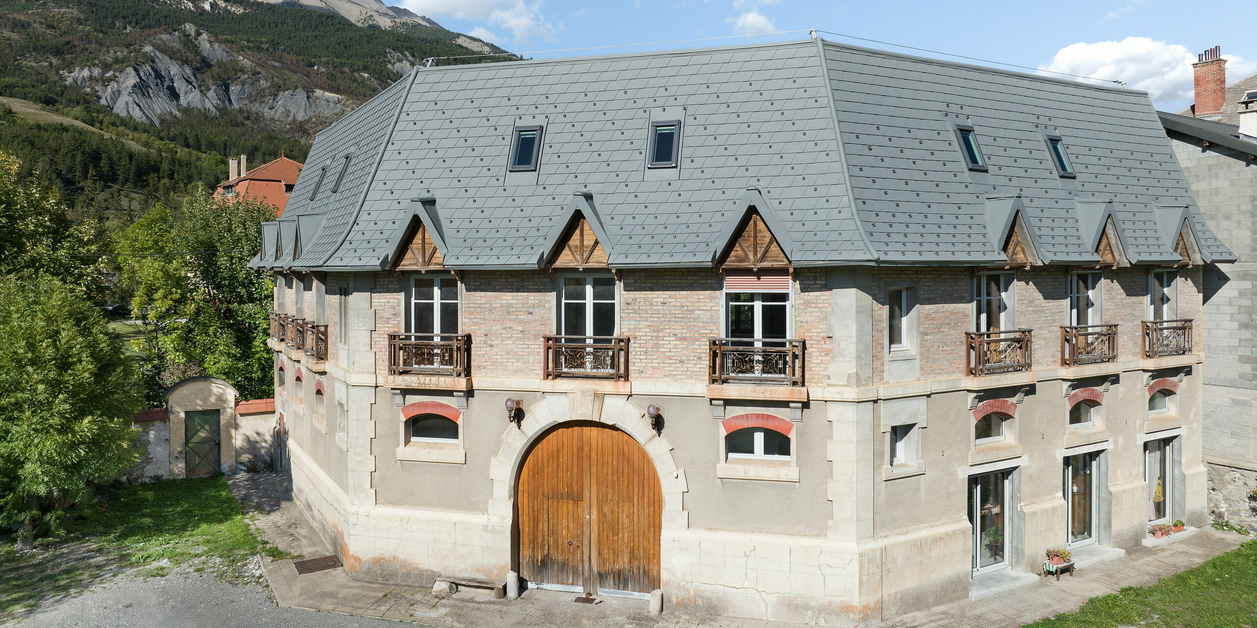 Frontansicht eines Anwesens in Barcelonnette, Frankreich. Das großflächige Dach wurde mit der PREFA Dachschindel in P.10 Hellgrau saniert. 