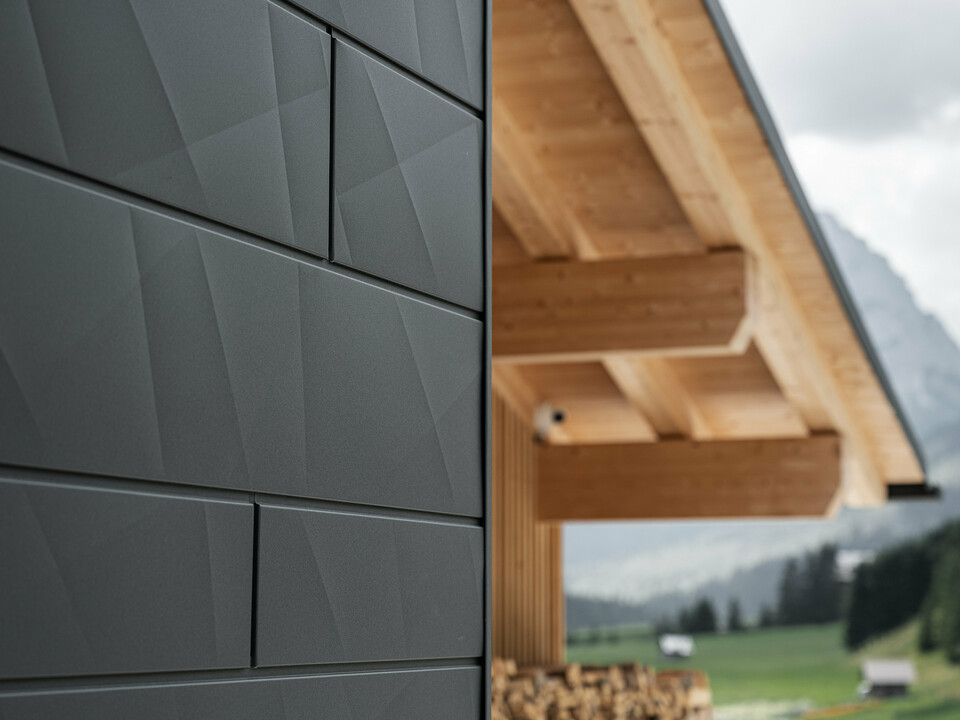 Nærbillede af Siding.X facadeplader i P.10 sort fra PREFA på Casa Hoffe i Sappada, Italien. Facadesystemet i aluminium er monteret i et moderne, abstrakt design, der danner en flot kontrast til de synlige træelementer i tagkonstruktionen. Den innovative facadeløsning passer perfekt ind i det alpine miljø og står for en kombination af funktionalitet, holdbarhed og æstetisk byggekultur.