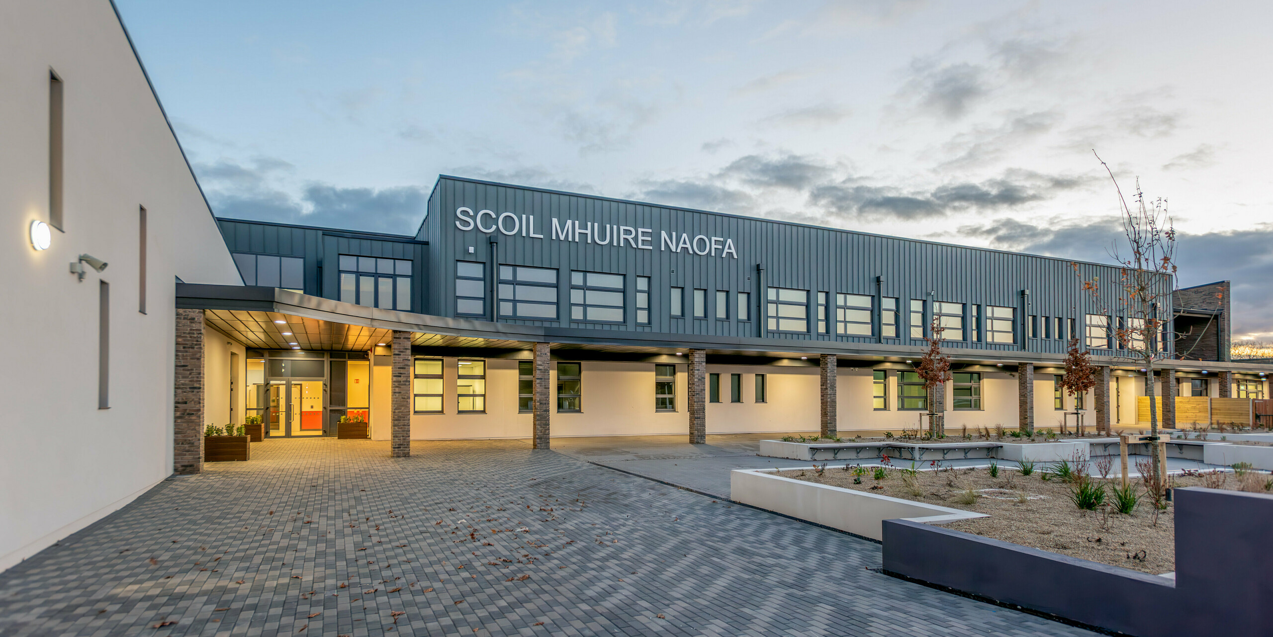 Die Scoil Mhuire Naofa im irischen Carrigtwohill in der Abenddämmerung. Die zeitgemäße Fassade aus PREFALZ Aluminium in der Farbe P.10 Hellgrau, kombiniert mit Backsteinsäulen und klaren Linien, verleiht dem Schulgebäude ein einladendes und hochwertiges Erscheinungsbild. Die klare Architektur, die stilvolle Beleuchtung und der gepflegte Vorplatz schaffen eine angenehme Atmosphäre.