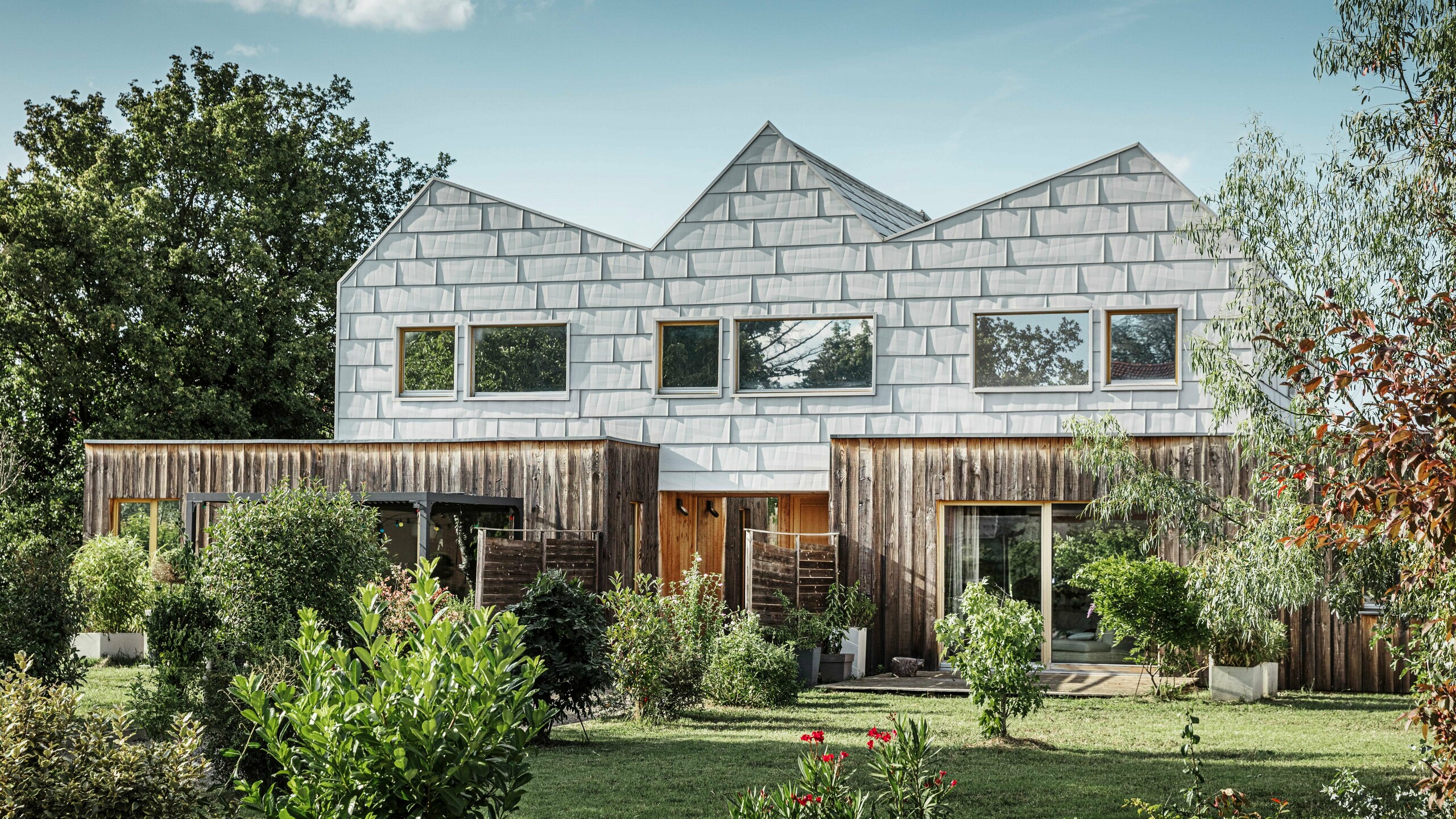 Modernes Doppelhaus mit Satteldach und PREFA-Fassade. Die Fassade besteht aus FX.12 Aluminiumpaneelen, die eine strukturierte, moderne Optik bieten. Das Haus ist von einem gepflegten Garten umgeben, und die Fensterrahmen harmonieren perfekt mit der Gesamtarchitektur.