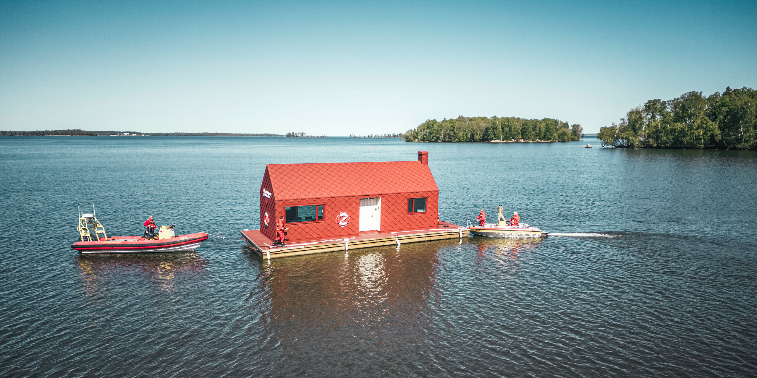 Luftaufnahme des Seenotrettungsstationshauses auf dem Hjälmaren See in Schweden, umgeben von der ruhigen Weite des Wassers. Das markante, oxydrote Gebäude, ausgestattet mit PREFA Dach- und Wandrauten 29×29, thront auf einem Schwimmkörper und ist bereit, den Rettungskräften Zuflucht und Betriebsbasis zu bieten. Neben dem Stationshaus, das sich durch seine charakteristische Farbgebung und Form hervorhebt, patrouillieren zwei Rettungsboote, welche die ständige Wachsamkeit und Einsatzbereitschaft der Seenotretter symbolisieren.