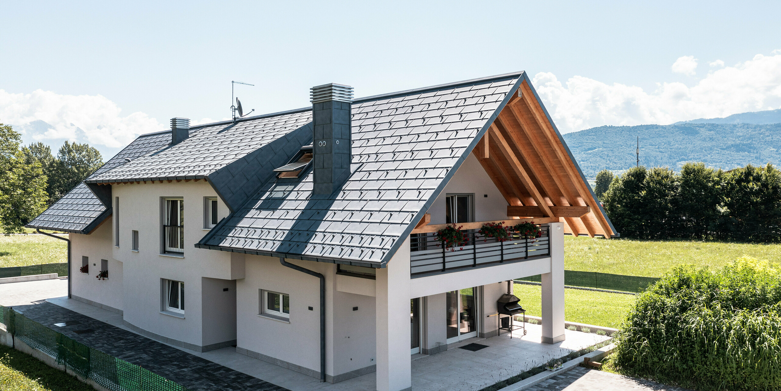 Charmantes Einfamilienhaus in Bellun, Norditalien, gekrönt mit einer Dachplatte PREFA R.16 in Anthrazit. Das Dach mit seiner P.10 Beschichtung spiegelt die robuste Eleganz und technische Reife wider, die PREFA Produkte auszeichnen. Die Farbe steht im Einklang mit den saftig grünen Wiesen und dem Bergpanorama im Hintergrund. Das Bild zeigt die Terrasse mit der darüber liegenden Loggia.