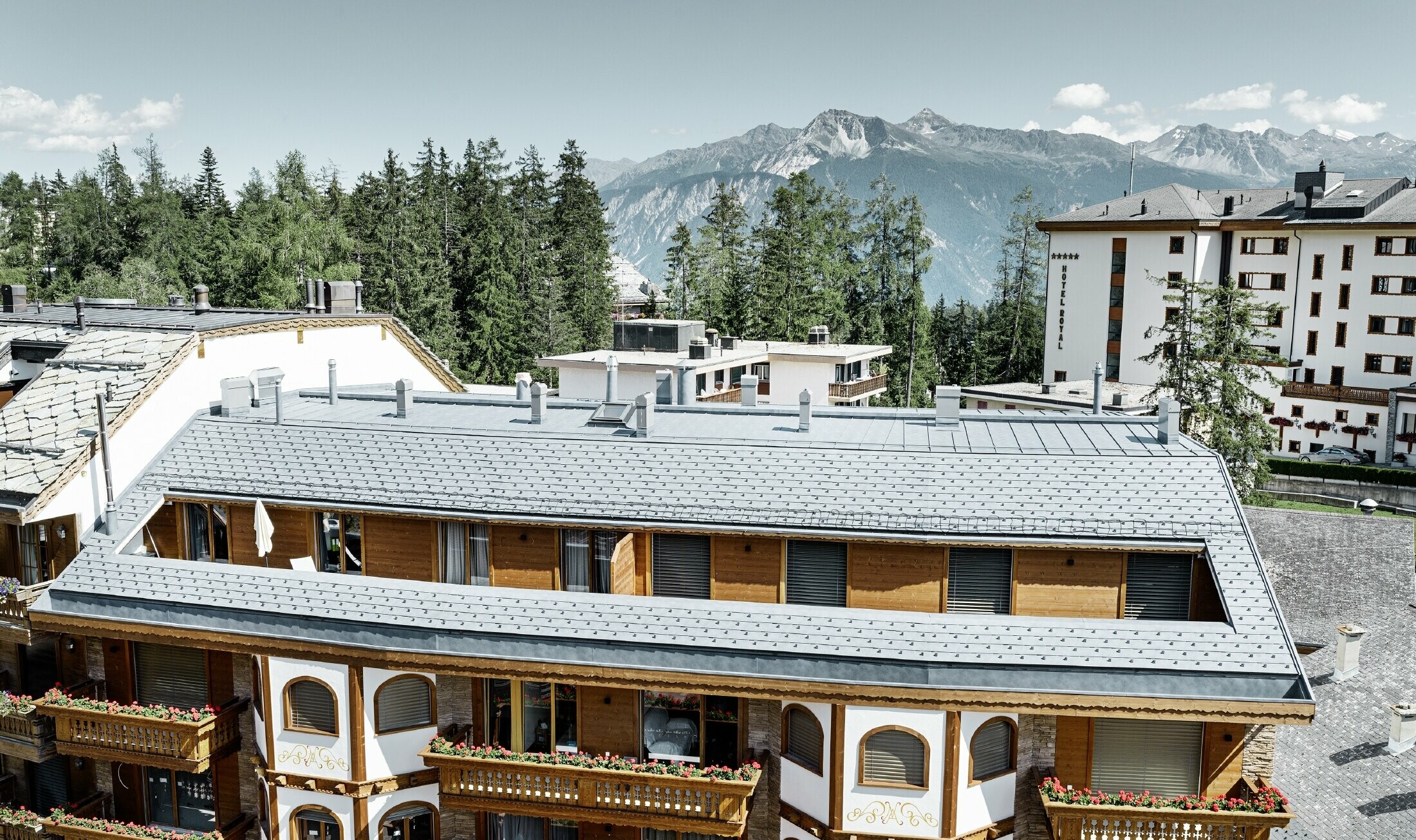 Flerfamiliehus i Crans-Montana med bjerge i baggrunden og en facade med fantasifulde træelementer med en PREFA aluminium tagspån-tag i stengrå