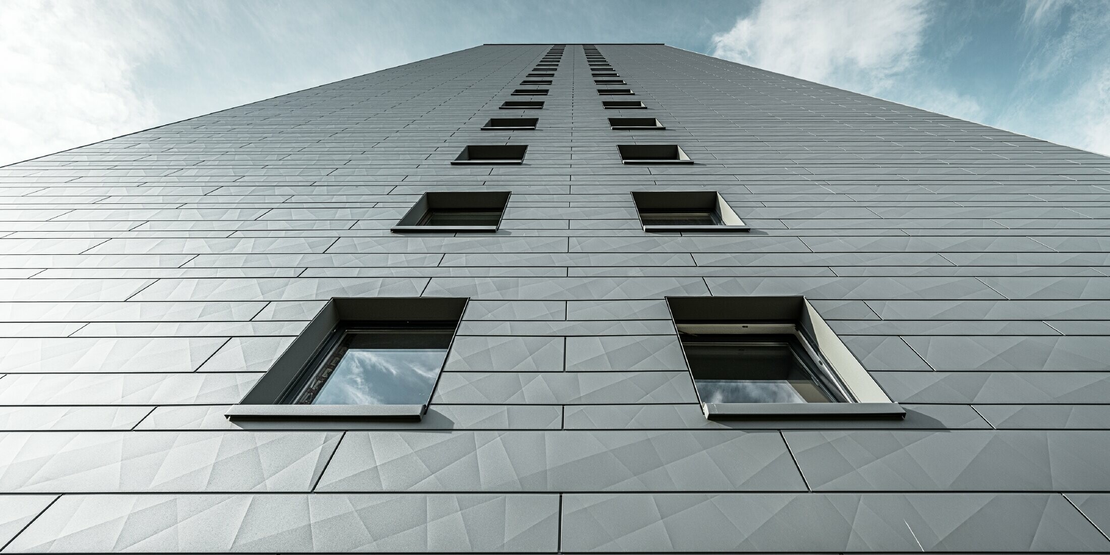 Blick an der Fassade hoch an einem vielstöckigen Hochhaus, die Fassade ist verkleidet mit waagrechten Siding.X Fassadenpaneelen von PREFA in Hellgrau.