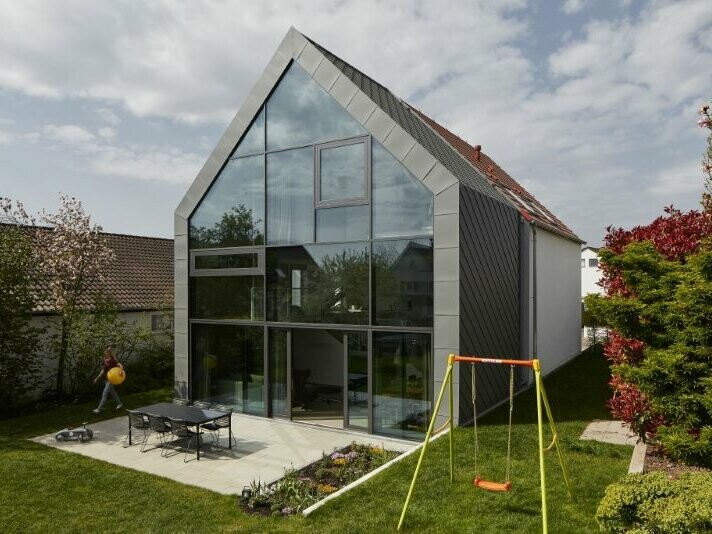 Vorderansicht des Haus J in Stuttgart (Deutschland). Im Hintergrund erkennt man einen bewölkten Himmel. Das Haus J wurde mit der Dachraute 29 × 29 in der Farbe P.10 Hellgrau verkleidet.