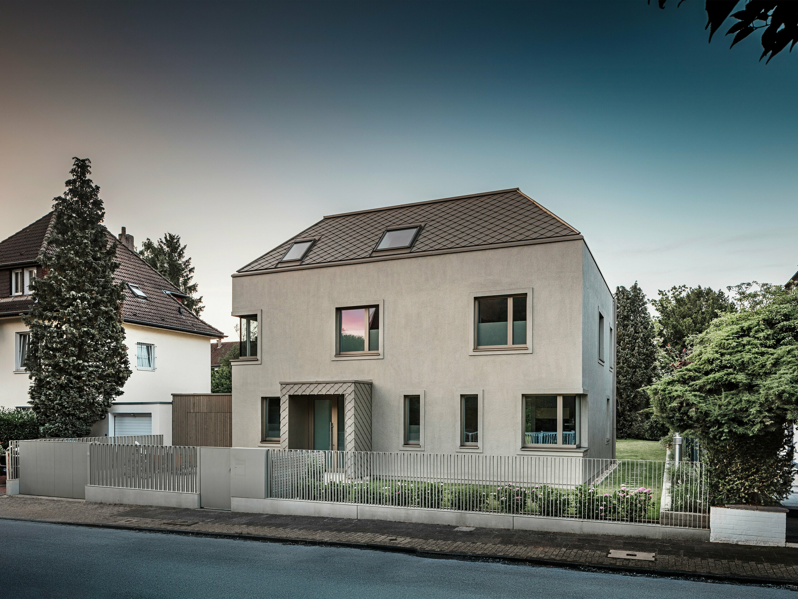 Ein modernes Einfamilienhaus mit Walmdach und bronze-farbener Aluminium-Dach- und Fassadenverkleidung von PREFA. Das Haus befindet sich in einer ruhigen Wohngegend, umgeben von gepflegten Gärten und anderen Einfamilienhäusern. Im Vordergrund ist ein niedriger Zaun zu sehen, der das Grundstück von der Straße trennt. Die Architektur des Hauses zeichnet sich durch klare Linien und große Fenster aus, die viel natürliches Licht in die Innenräume lassen. 