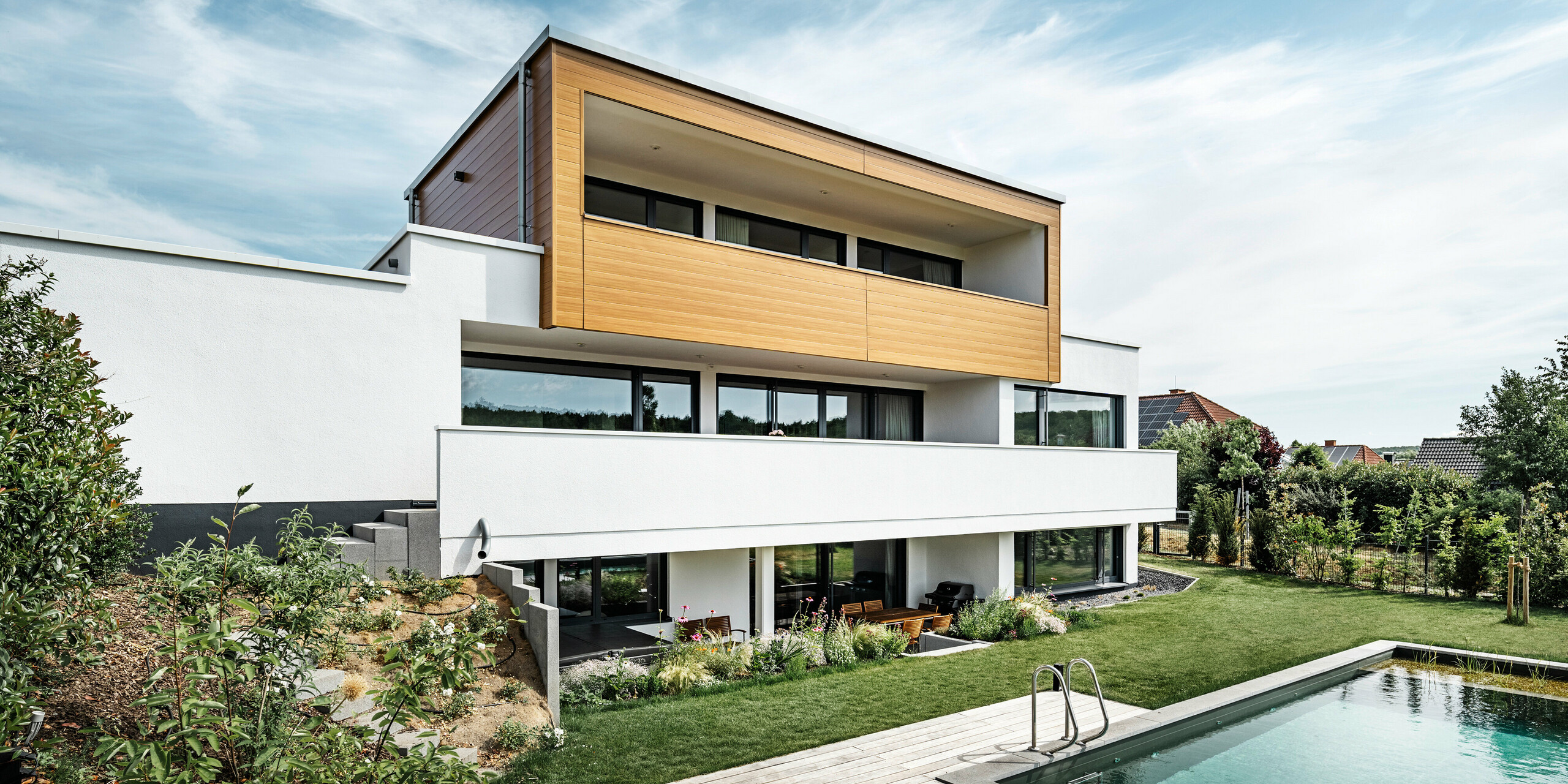 Rückseite eines Einfamilienhauses in Usingen. Zu sehen ist der schöne Garten inkl. Pool sowie die markante Fassade bestehend aus einem weißen Gebäudesockel und PREFA Sidings in Eiche natur am Obergeschoss.