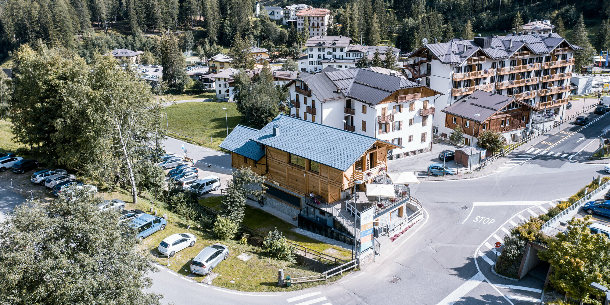 Vogelperspektive auf eine belebte Kreuzung in einer alpinen Dorfgemeinschaft mit einem hervorstechenden Après-Ski-Lokal mit PREFA Dachschindeln DS.19 in P.10 Steingrau. Die Kombination aus traditioneller Holzbauweise und modernen Dach-Elementen aus Aluminium, umgeben von einer Kulisse aus dicht bewaldeten Hügeln und einer Mischung aus modernen und traditionellen Gebäuden, zeugt von einer gelungenen Symbiose aus Innovation und Tradition im Herzen der italienischen Alpen.