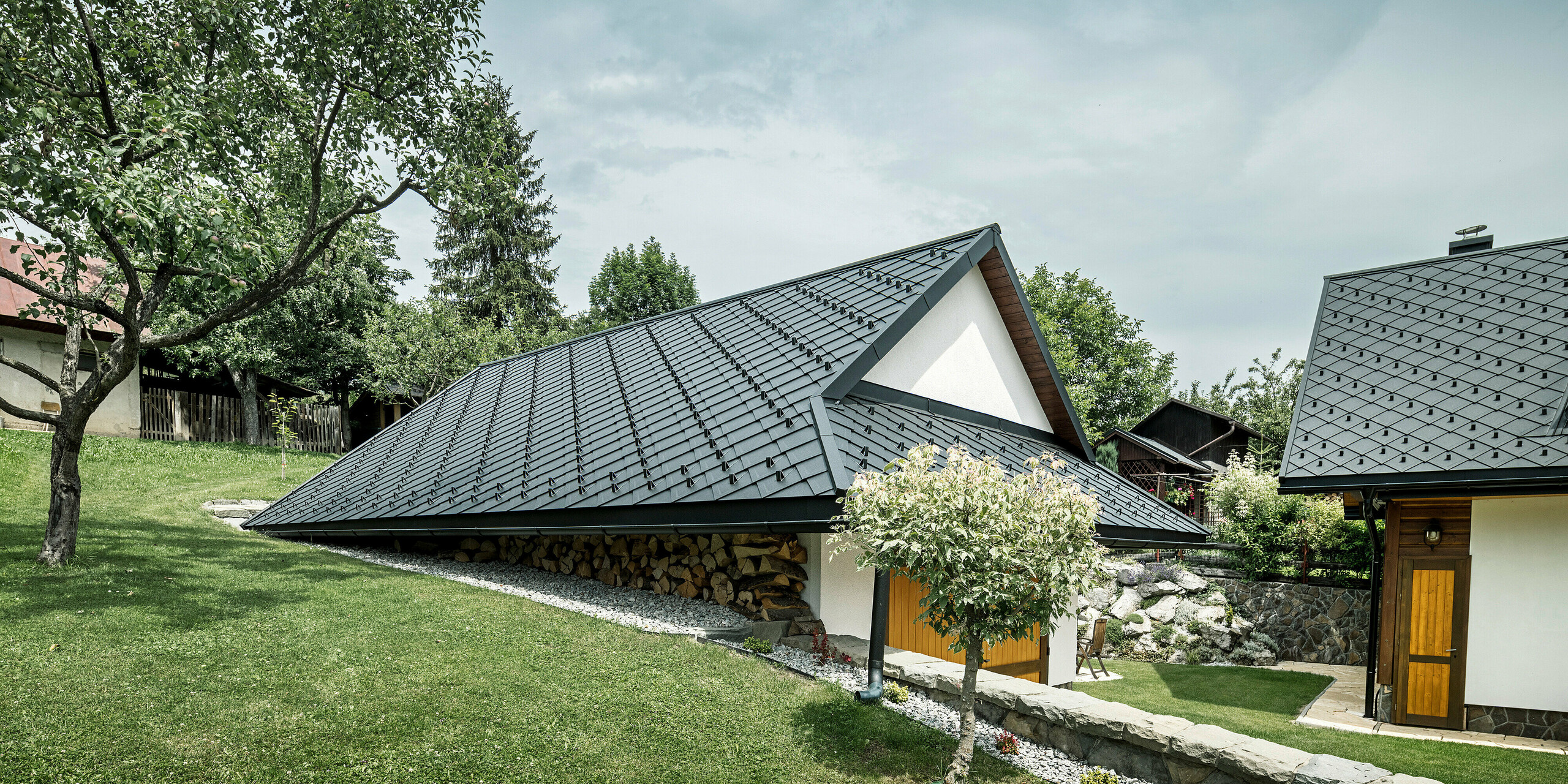 Nebengebäude eines traditionellen Einfamilienhauses in Stara Bystrica, Slowakei, mit einem Dach aus PREFA Dachrauten 29 × 29 in P.10 Schwarz. Das robuste Aluminiumdach in Schuppenoptik schützt das Gebäude und passt sich harmonisch in die grüne, ländliche Umgebung ein. Im Vordergrund ist eine gepflegte Rasenfläche und ein Baum zu sehen, was dem Gesamtbild eine idyllische und einladende Atmosphäre verleiht.