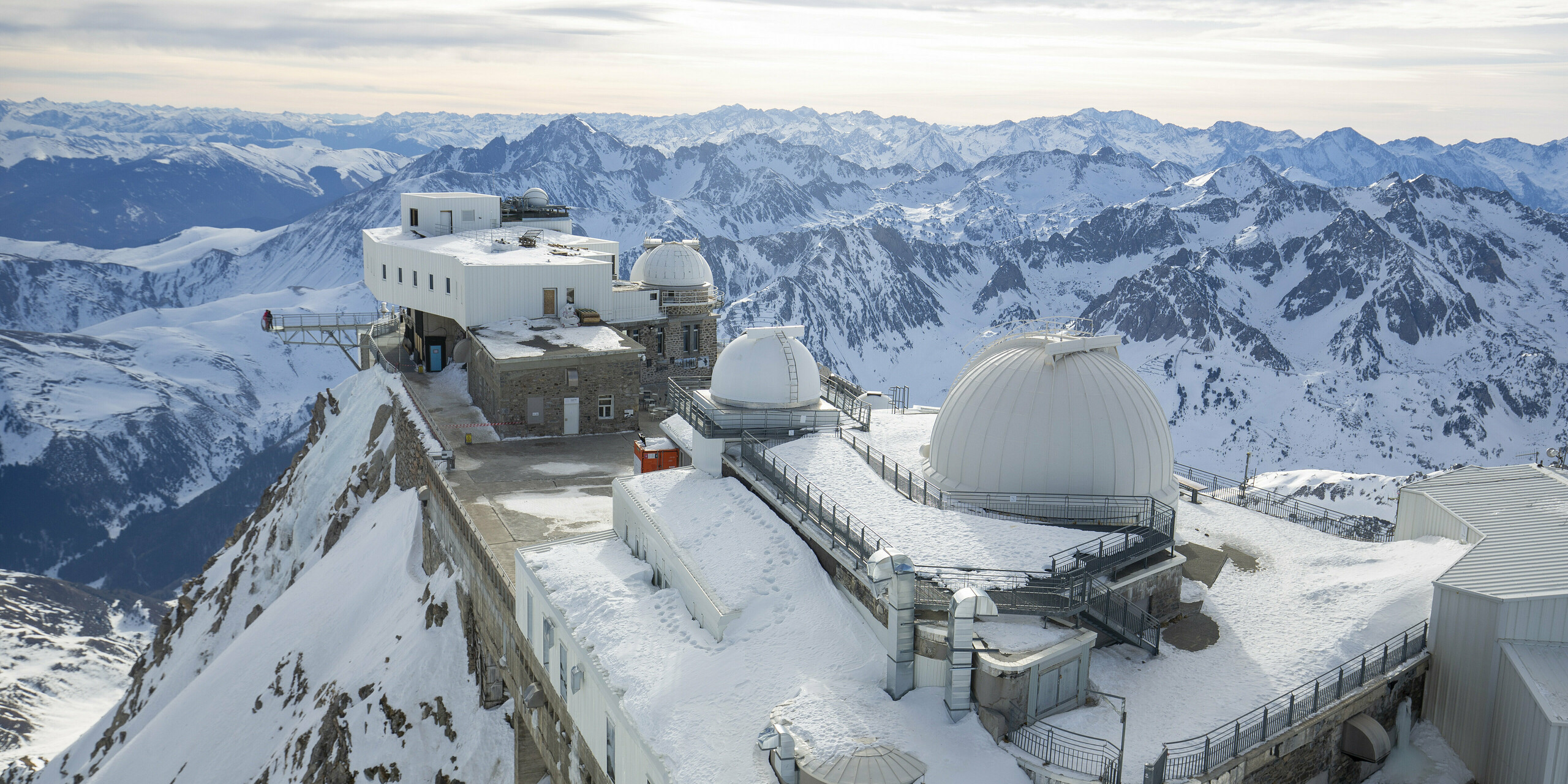 Das Midi-Pyrénées-Observatorium, auf einem verschneiten Berggipfel gelegen, zeigt seine umfassend sanierte und erweiterte Struktur. Die Fassade des Hauptgebäudes ist mit PREFALZ in der Farbe P.10 Reinweiß verkleidet, was aufgrund der extremen Hochgebirgsbedingungen ideal ist. Mehrere Observatoriumskuppeln und die modernen Anbauten sind sichtbar. Die umliegenden, schneebedeckten Pyrenäen erstrecken sich majestätisch im Hintergrund. Das robuste PREFALZ Dach- und Fassadensystem überzeugt durch unterschiedliche Eigenschaften. Unter anderem konnte die extreme Robustheit und das geringe Gewicht überzeugen