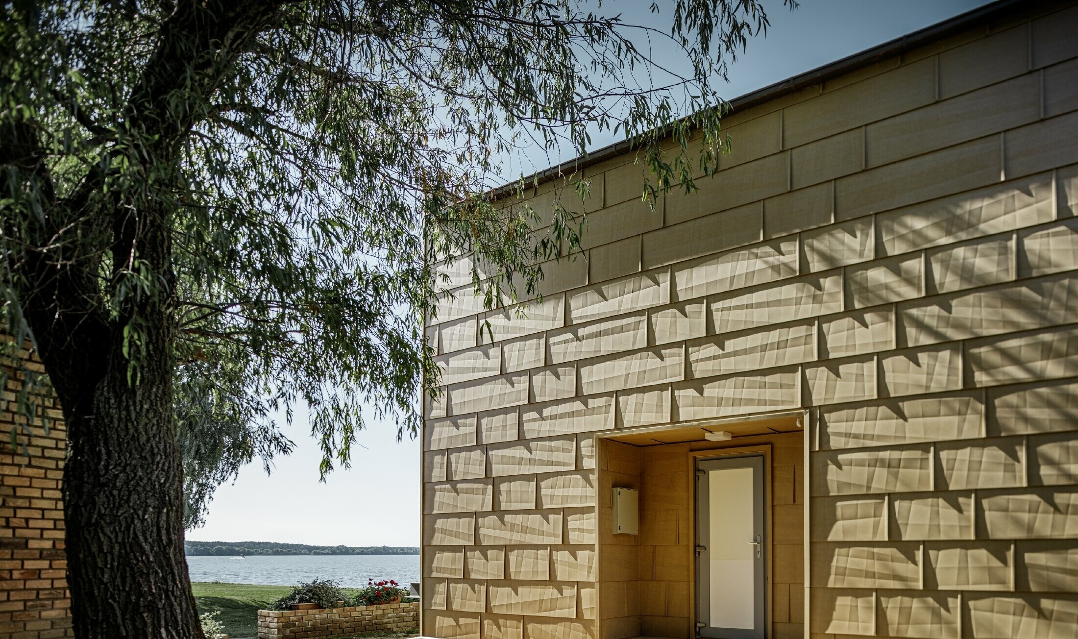 Moderne weekendhus med fladt tag og store vinduesflader ved havet med en vinklet aluminiumfacade i sandbrun