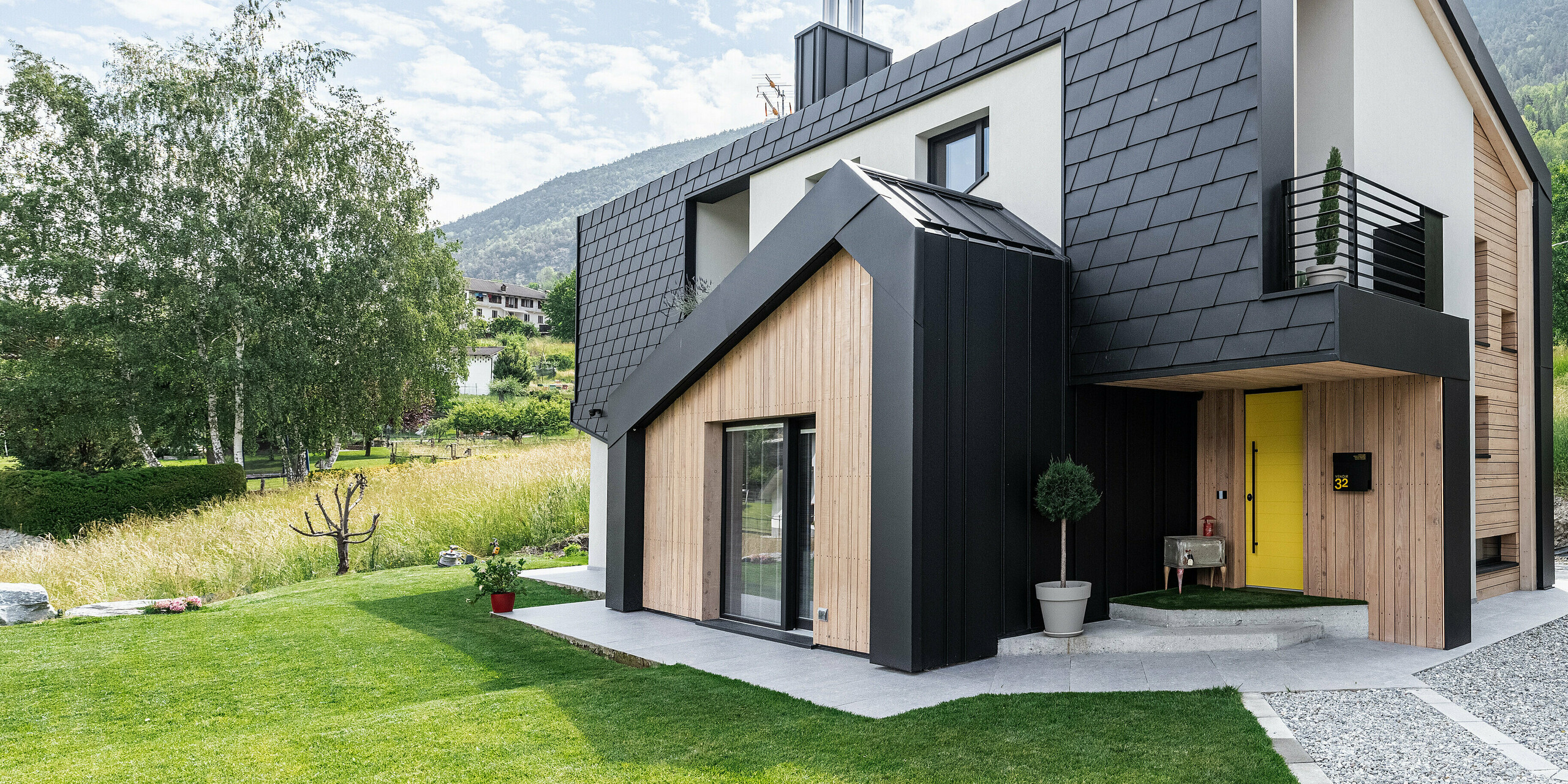 Elegantes Einfamilienhaus mit PREFA Dachschindeln DS.19 und Wandschindeln in P.10 Schwarz, umgeben von einer gepflegten Rasenfläche und einer natürlichen Landschaft. Das Haus zeichnet sich durch seine moderne Bauweise mit klaren Linien, Holzakzenten und einer auffälligen gelben Haustür aus, die einen einladenden Kontrast zur dunklen Gebäudehülle aus Aluminium bildet.