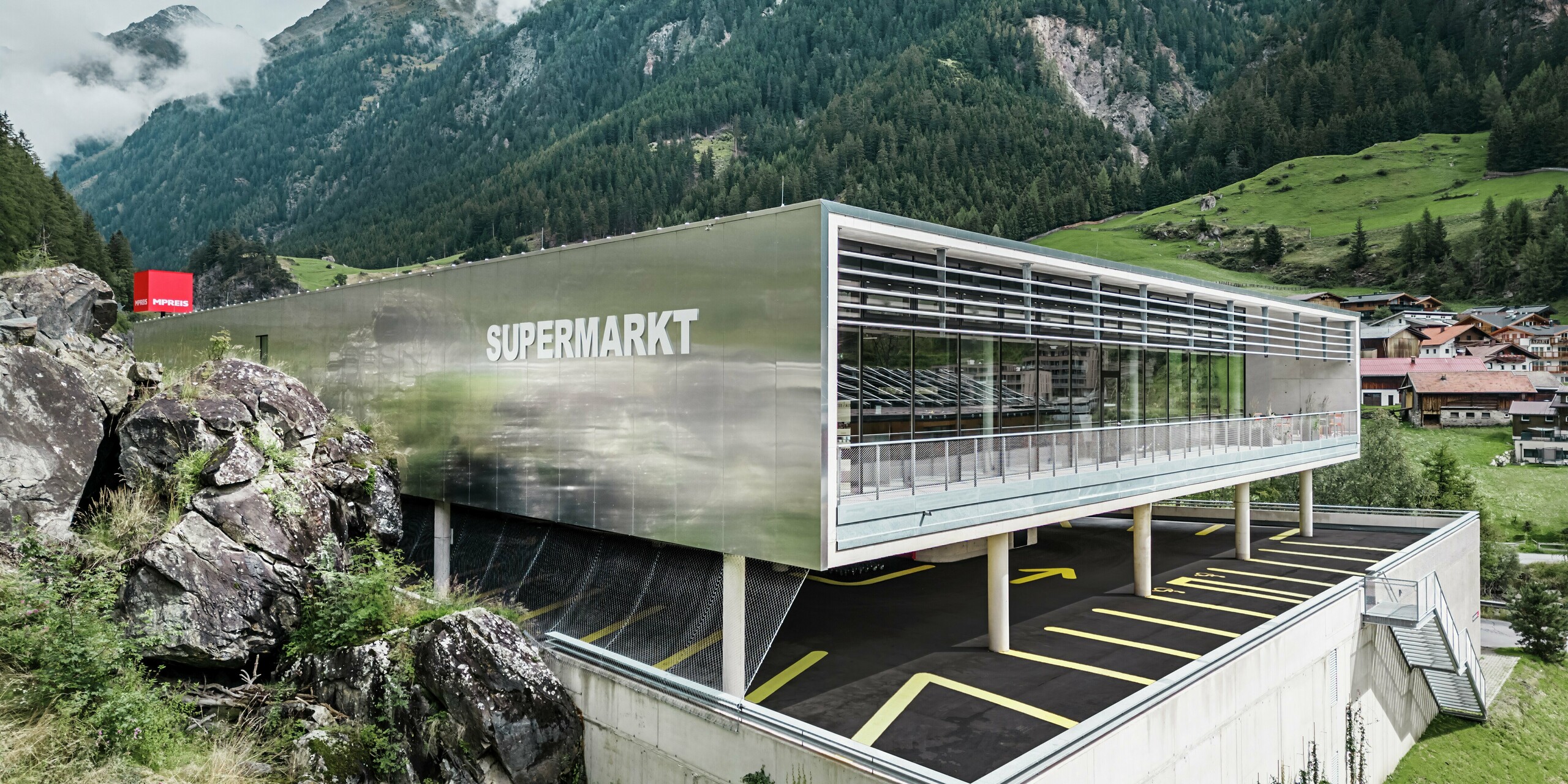 MPREIS-Markt in Sölden, Tirol, mit Aluminium Verbundplatten in der Sonderfarbe Reflective Natural. Die Fassade spiegelt die umliegende Berglandschaft der Ötztaler Alpen wider und verbindet modernes Design mit der Natur. Die klare Architektur mit dem Parkdeck unter dem Supermarkt schafft eine innovative Einkaufsmöglichkeit für den alpinen Raum.