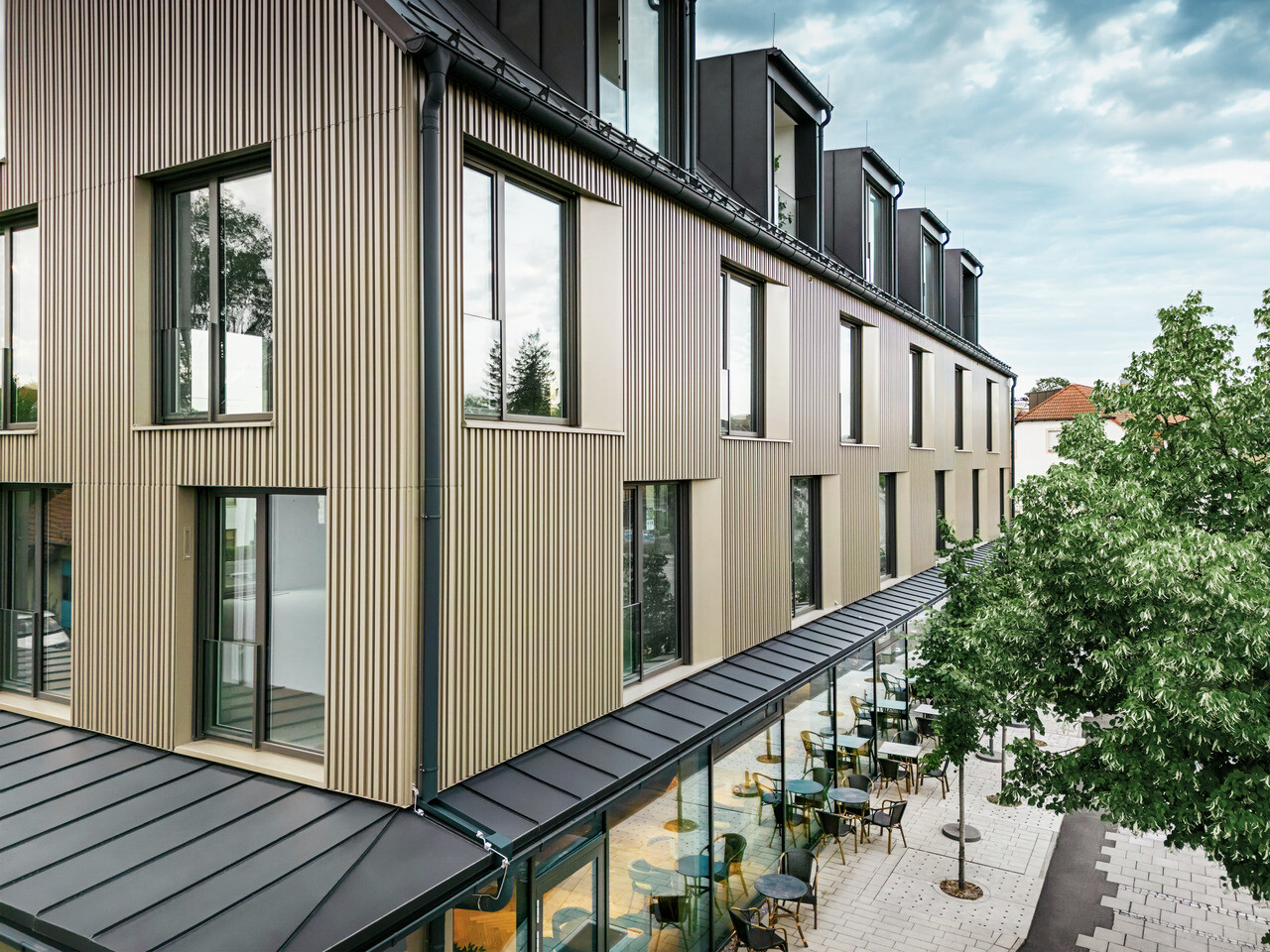 Das Bild zeigt ein modernes Gebäude in Feldkirch, entworfen vom Architekten Stefan Heigl. Die Fassade ist aus Prefalz in der Farbe Bronze gestaltet, und das Satteldach ist mit Prefalz in Schwarzgrau gedeckt. Große Fenster sorgen für viel Licht im Inneren, während die verglaste Erdgeschosszone eine einladende Gastronomiefläche bietet. Die hochwertige Materialwahl und das elegante Design verleihen dem Gebäude ein zeitgemäßes und stilvolles Erscheinungsbild.