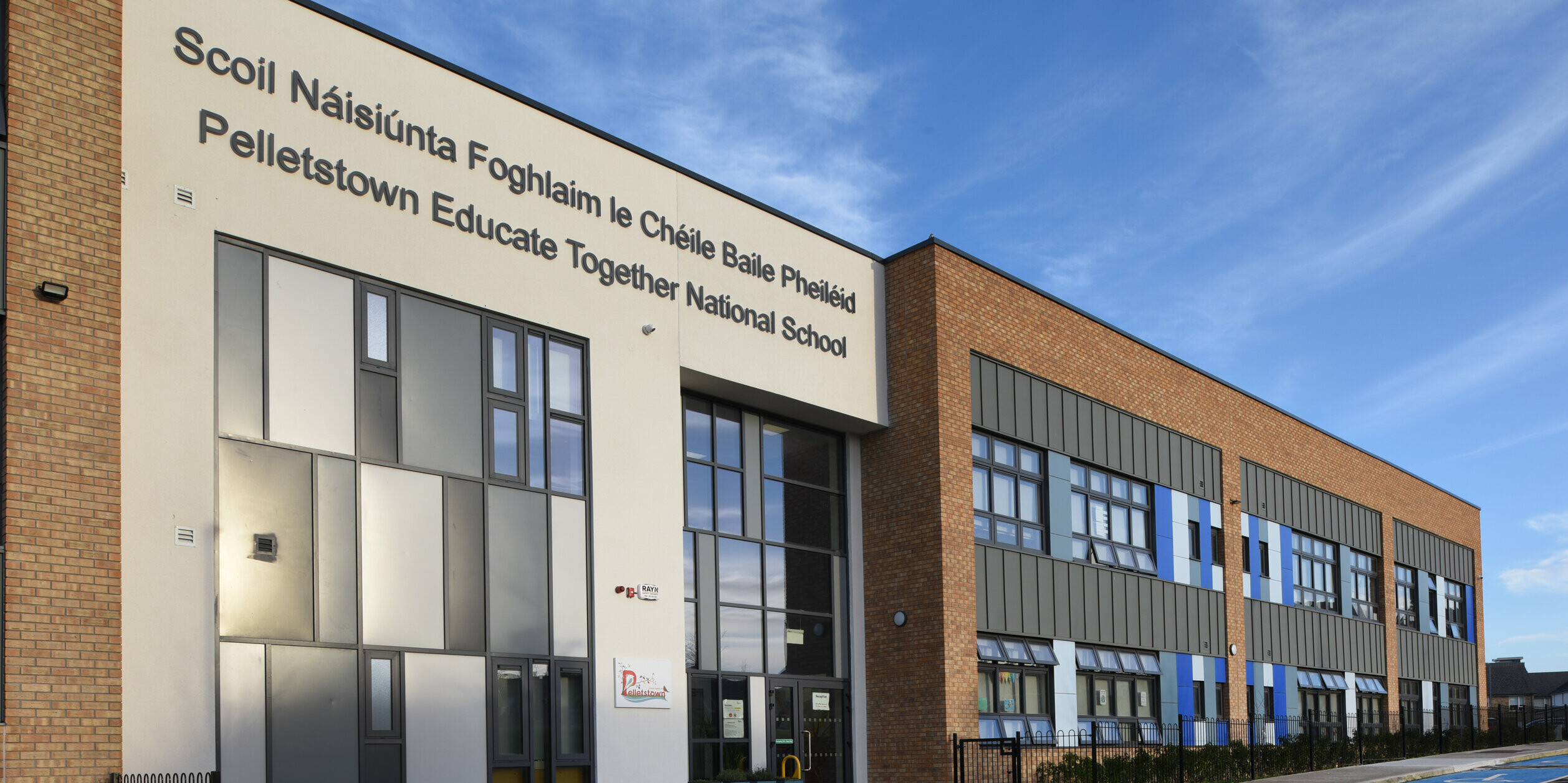 Eingangsbereich der Pelletstown Educate Together National School in Dublin. Die Fassade der Schule besteht aus einem spannenden Materialmix aus Aluminium, Putz und traditionellen Ziegeln. Die Stehfalzdeckung aus PREFALZ in P.10 Hellgrau unterstreicht die moderne und nachhaltige Architektur. Fahrräder vor dem Eingang zeigen, dass die Schule großen Wert auf Nachhaltigkeit legt. Unter den Fensterreihen sorgen Sträucher für eine optische Auflockerung der robusten Baumaterialien.