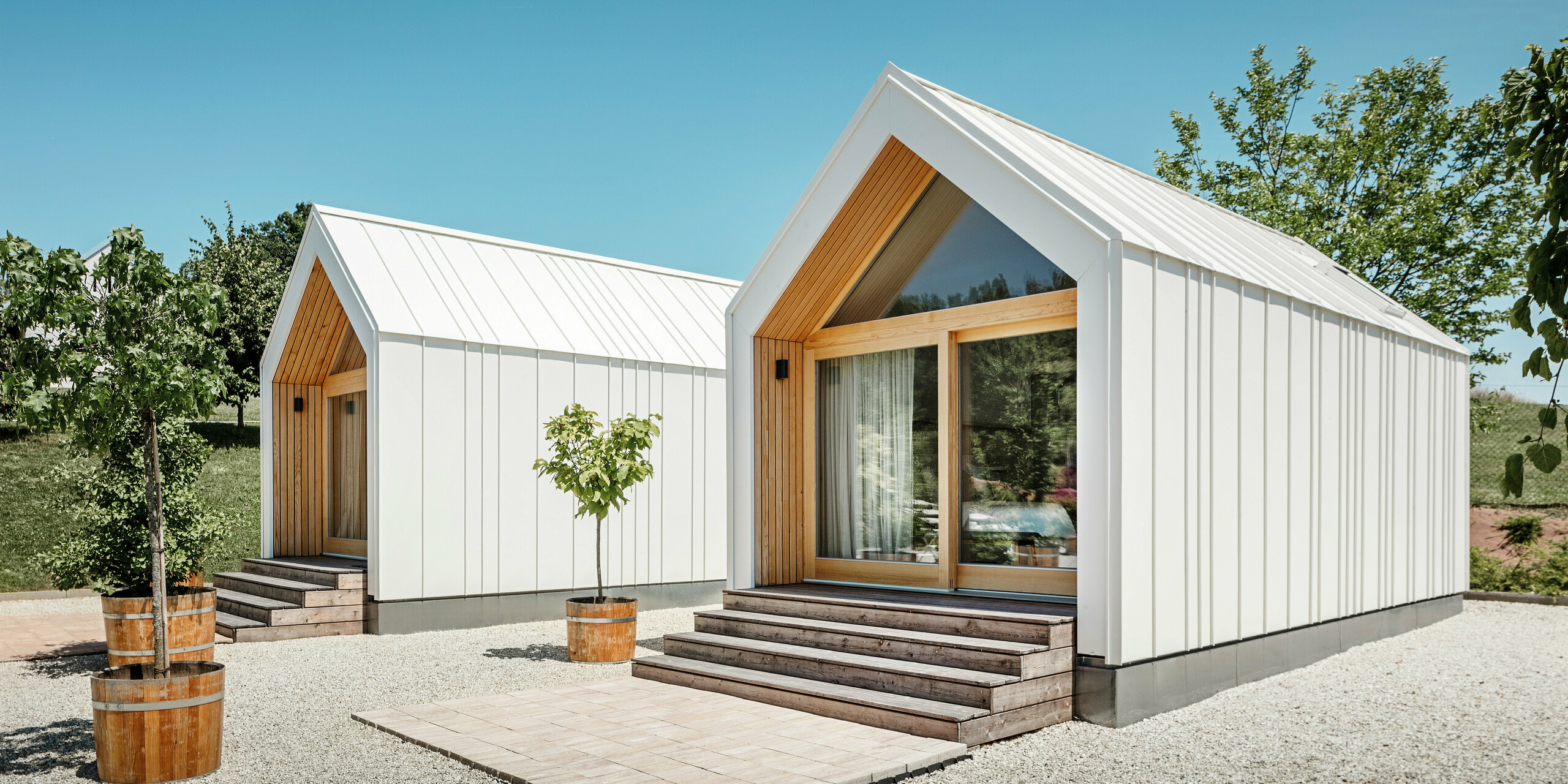 Zwei weiße Tiny Häuser der Ferienidylle 'Pri Momi' in Kančevci, Slowenien, gestaltet mit PREFALZ Dach- und Fassadenverkleidung in P.10 Reinweiß und naturbelassenem Holz. Die minimalistische Architektur der Gebäude wird durch den eleganten Materialmix aus Aluminium und Holz unterstrichen. Die großen Glasfronten an den Eingängen sowie die Holzstufen und umliegenden Bäume schaffen eine einladende und harmonische Atmosphäre. Die exklusive Unterkunft liegt inmitten einer malerischen Landschaft und wird speziell für Feiern genutzt.