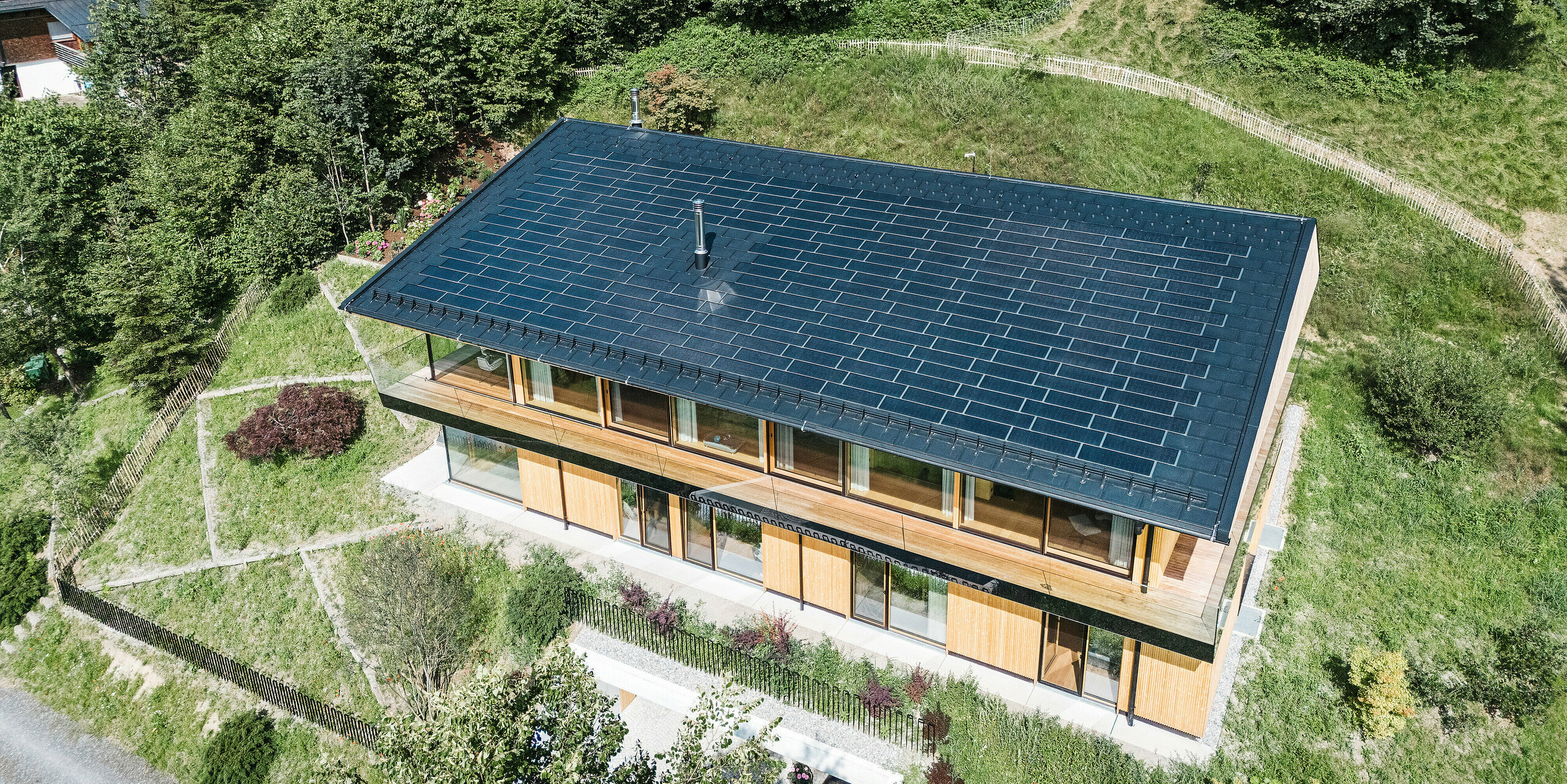 Vogelperspektive auf ein Einfamilienhaus in Egg, Vorarlberg, mit einem Dach aus PREFA Solarplatten in P.10 Schwarz. Das Aluminiumdach mit integrierter Photovoltaikanlage ermöglicht die Stromerzeugung aus erneuerbarer Energie, ohne die homogene Einheit des Daches zu beeinträchtigen. Die großzügige Glasfassade und die Holzverschalung harmonieren perfekt mit der umgebenden Natur.