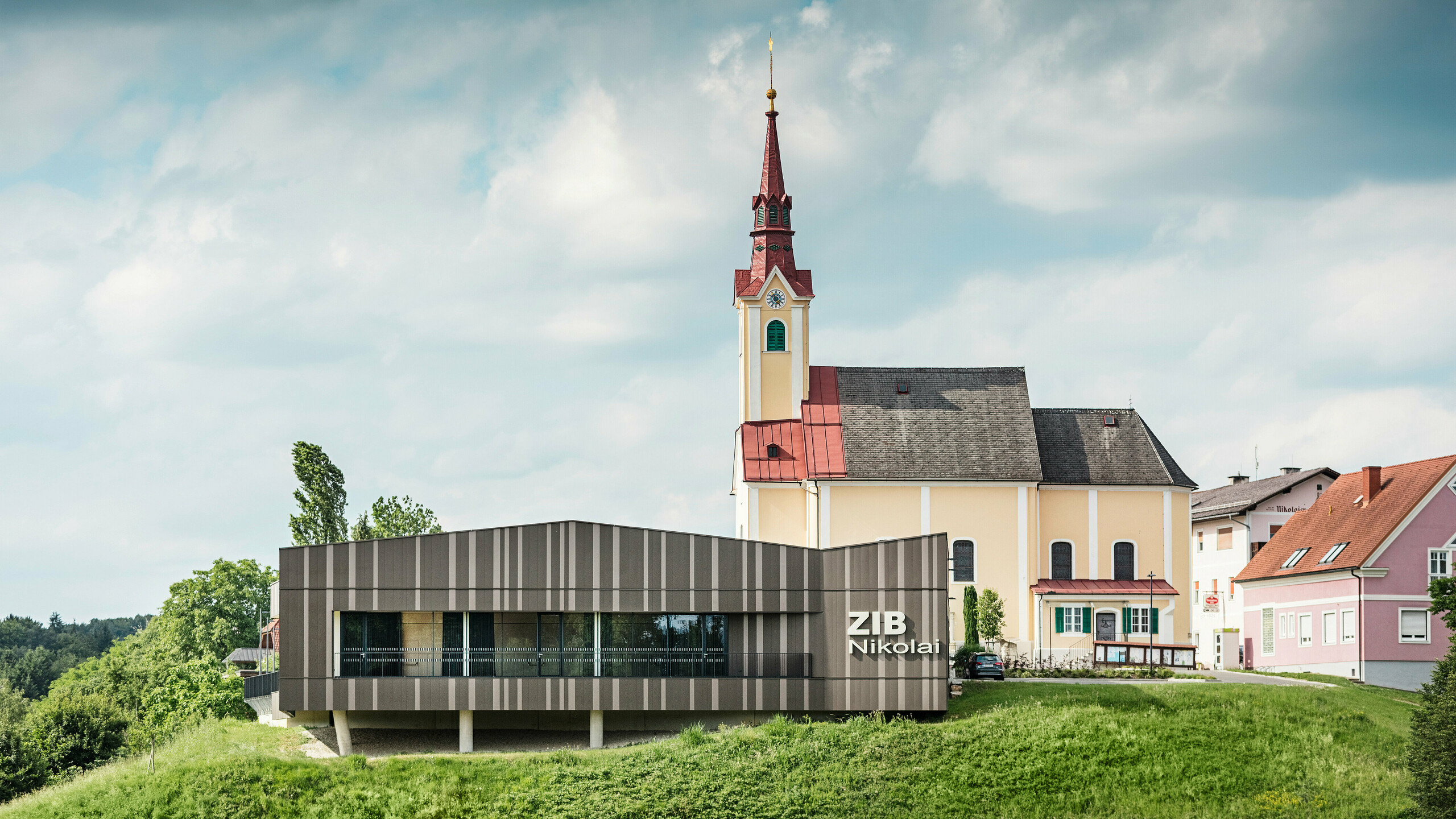 Das Veranstaltungszentrum ZIB Nikolai in St. Veit in der Südsteiermark mit einer modernen, dynamischen Aluminiumfassade in braun-beigegrauen Tönen, im Vordergrund eine grüne Wiese, im Hintergrund die Kirche von St. Nikolai mit gelber Fassade und rotem Dach.