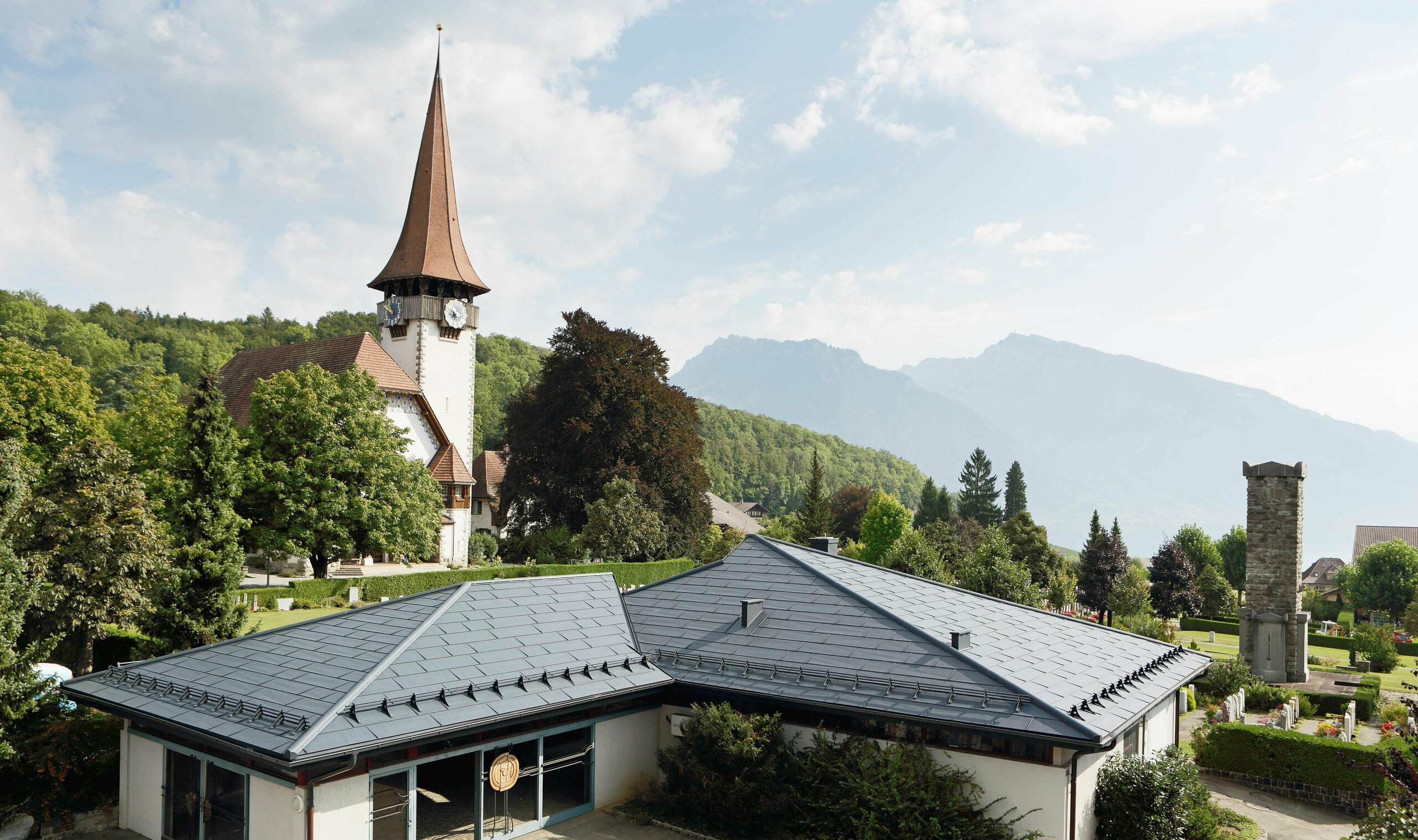 Kapel med FX.12-tagpaneler fra PREFA i antracit; i baggrunden ses en kirke og ved siden af den kirkegården.