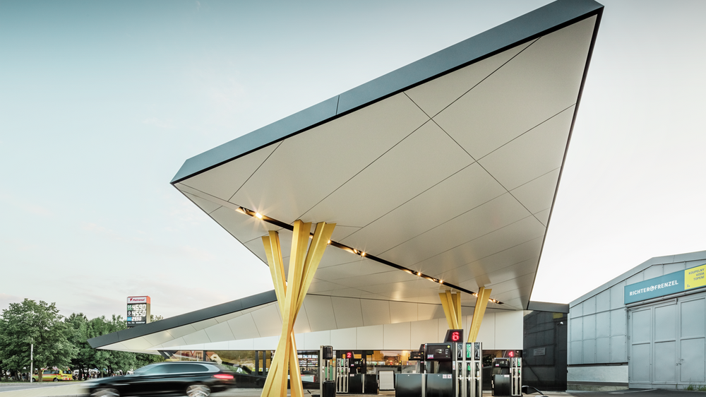 Seitliche Ansicht der Tankstelle mit einer Untersicht des Daches, welches auf zwei goldenen Stützengruppen aufliegt.