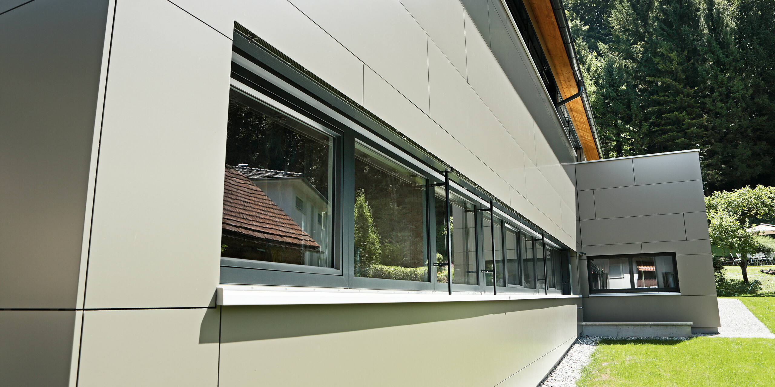 Nahaufnahme des Erdgeschosses eines Einfamilienhauses in Hohenems, das mit PREFA Aluminium Verbundplatten in der Farbe Bronzemetallic verkleidet ist. Die glatten, sauber verarbeiteten Fassadenelemente bieten eine elegante Optik und unterstreichen die großen Fensterpartien, die das Tageslicht maximieren. Die Detailaufnahme zeigt die präzise Handwerkskunst und Qualität der Materialien, die das Haus mit einem zeitgemäßen und langlebigen Äußeren versehen.