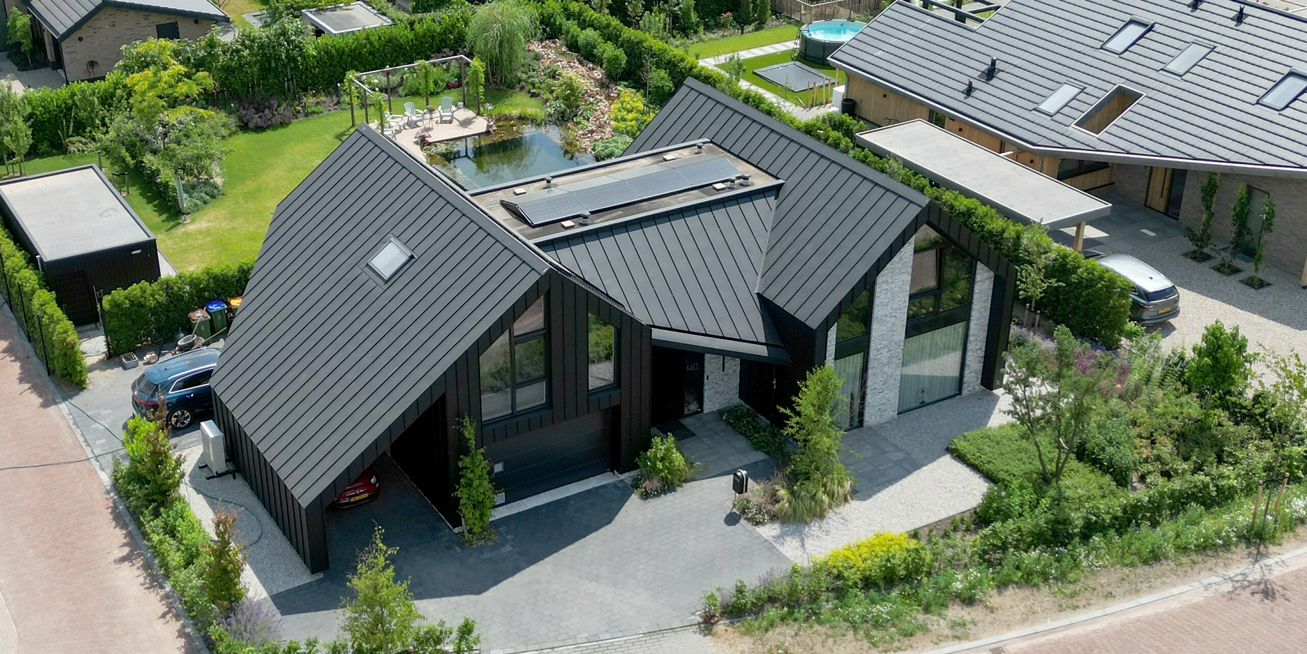 Blick aus der Vogelperspektive auf ein beeindruckendes Einfamilienhaus in Veenendaal, Niederlande, das sich durch sein schwarzes PREFALZ Dach auszeichnet und sich harmonisch in die umgebende Gartenlandschaft einfügt. Das Haus hat ein modernes Satteldach und ist dank seiner klaren Linien und großen Glasflächen, die von lebendigem Grün umrahmt sind, ein Paradebeispiel für moderne Architektur. Die gut durchdachte Gartengestaltung mit privater Auffahrt und Garten unterstreicht die durchdachte Ästhetik des Hauses.