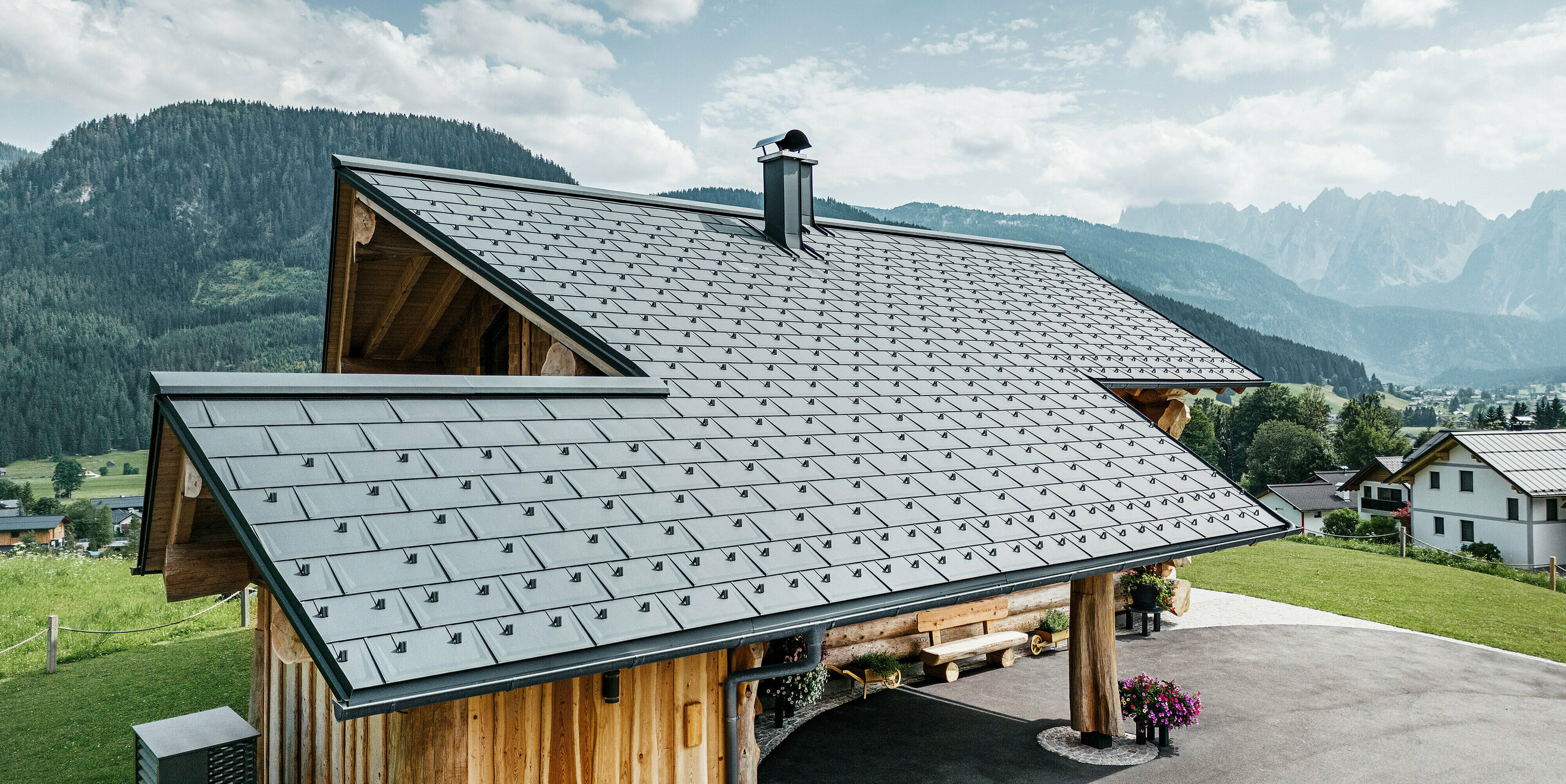 Moderne und zugleich traditionelle Architektur vereint in einem Blockhaus in Gosau, Österreich. Das Holzhaus verfügt über ein Dach aus PREFA Dachplatten R.16 in P.10 Anthrazit. Die Luftaufnahme zeigt die Präzision des Dachaufbaus, die perfekte Integration in das Blockhausdesign und die idyllische Einbettung in die alpine Landschaft mit einem atemberaubenden Bergpanorama im Hintergrund. Die Dachplatten verleihen dem Haus eine zeitgemäße Note und stehen im Einklang mit einer umweltfreundlichen und nachhaltigen Bauweise.