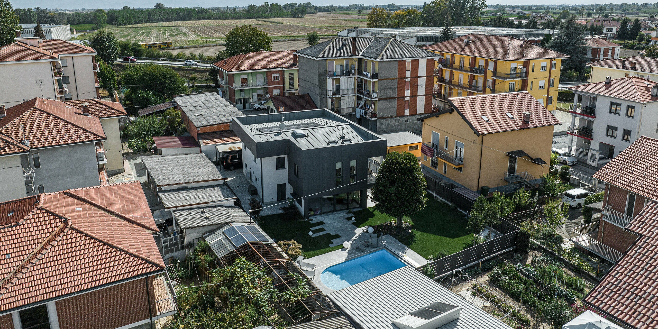 Luftaufnahme eines modernen Einfamilienhauses in Savigliano, Italien, mit einer dunkelgrauen PREFA Siding.X Aluminiumfassade. Diese Vogelperspektive zeigt das Haus eingebettet in eine kleinstädtische Umgebung mit umliegenden traditionellen Gebäuden und einem klaren Blick auf die fernen Berge. Die elegante und dezente Fassade des Hauses sticht in der bunten Nachbarschaft hervor, während der gut gepflegte Garten mit Pool eine Oase der Entspannung bietet.