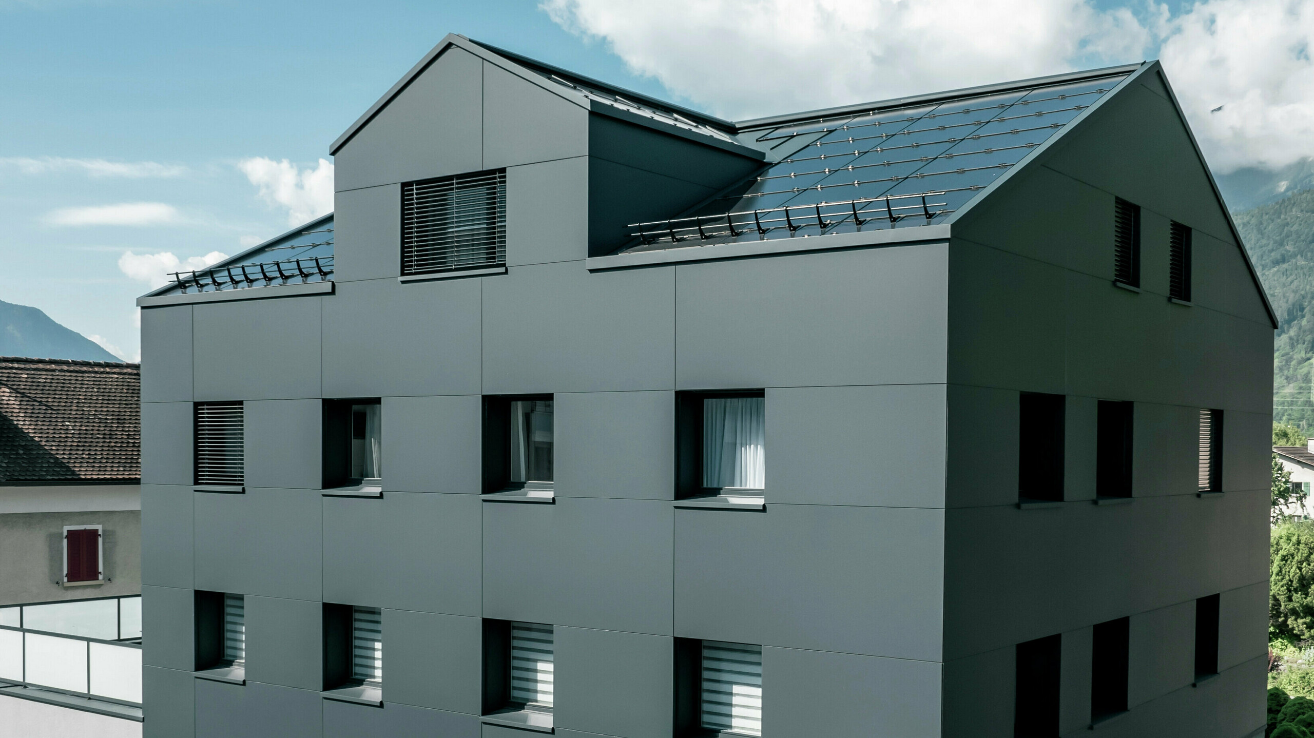 Modernisiertes Wohn- und Geschäftsgebäude nach der Fassadensanierung mit PREFABOND Aluminium Verbundplatten in Anthrazit. Die klare, elegante Linienführung der neuen Fassade verleiht dem Gebäude ein modernes Erscheinungsbild. Im Erdgeschoss befindet sich das Architekturbüro Voser. Die Umgebung mit Bäumen und Bergen rundet die harmonische Optik ab.