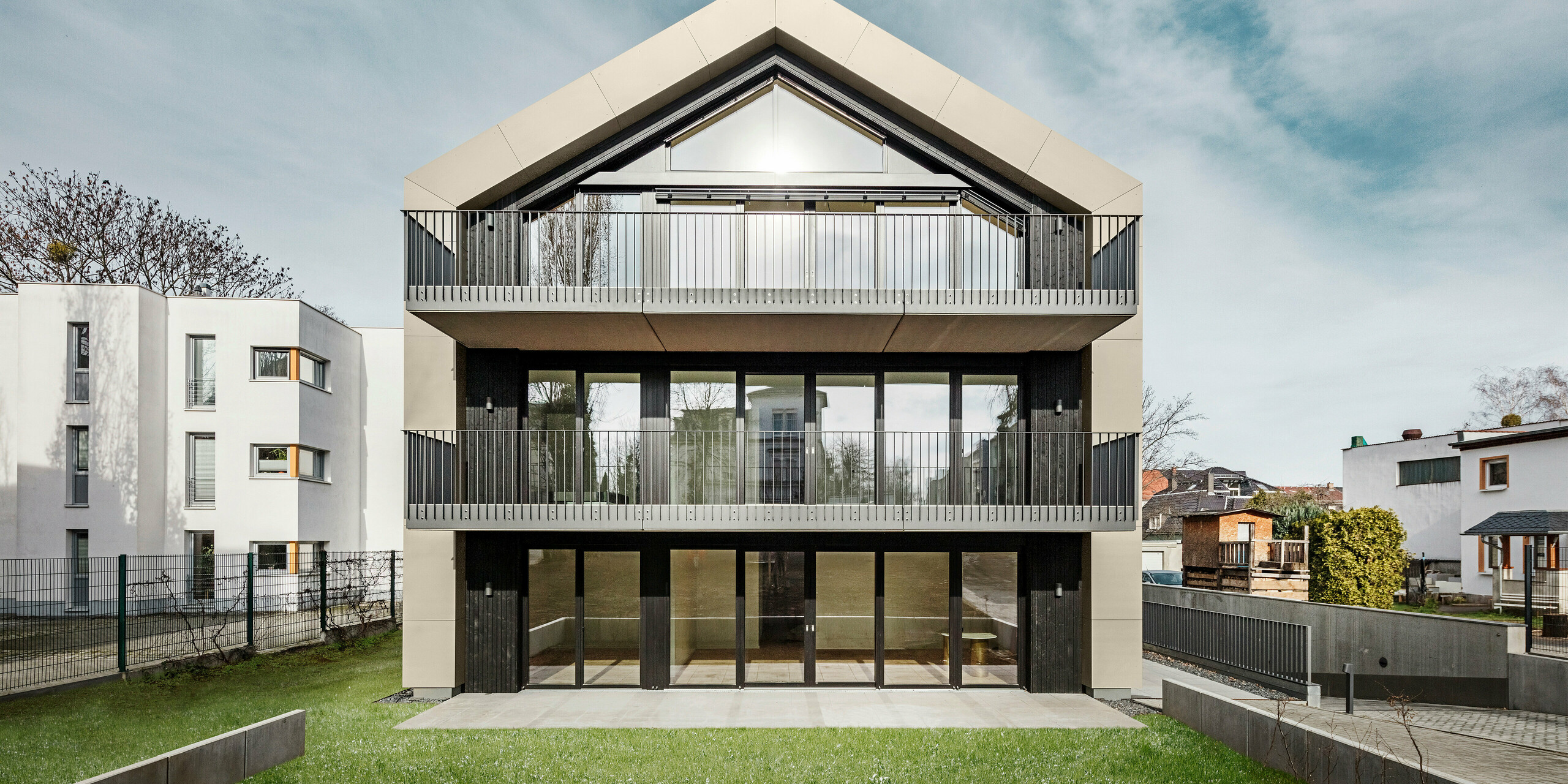 Frontalansicht eines modernen Mehrfamilienhauses in Dresden (Chemnitzer Straße) mit einer imposanten Glasfront und Balkonen. Die Fassade und das Dach des Gebäudes sind mit der langlebigen PREFA Aluminiumraute 29 × 29 in Bronze verkleidet, was dem Gebäude eine elegante und zeitgemäße Optik verleiht. Das Bild zeigt die symmetrische Gestaltung der Fassade, die großzügigen Glasflächen und den gepflegten Garten im Vordergrund.