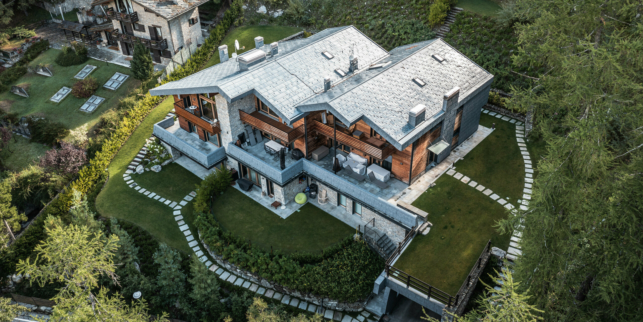 Eine Luftaufnahme einer beeindruckenden Villa in Courmayeur, die in eine üppige Landschaft eingebettet ist und eine perfekte Balance zwischen moderner und traditioneller Architektur zeigt. Die Kombination aus PREFA Aluminium in Form von FX.12 Dach- und Fassadenpaneelen in Steingrau, Holz- und Steinelementen in der Fassade schafft ein harmonisches Gesamtbild. Der sorgfältig angelegte Garten mit stufenförmig angelegten Grünflächen und steinernen Gehwegen sorgt für eine angenehme Außenumgebung, die die natürliche Schönheit der alpinen Landschaft hervorhebt.