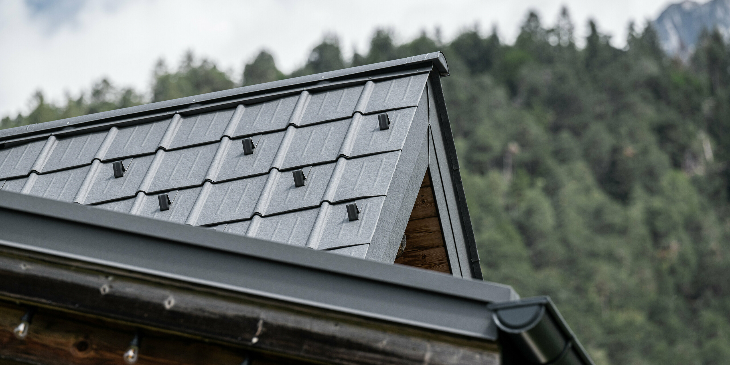 Nahaufnahme der Ecke eines traditionellen Alpenhauses mit modernen PREFA Dachplatten in P.10 Hellgrau, die eine ästhetische und wetterfeste Dachlösung darstellen Auronzo di Cadore, Italien. Die weichen Linien der Aluminiumziegel unterstreichen die hohe handwerkliche Qualität und das Engagement für eine harmonische Integration in die alpine Landschaft.