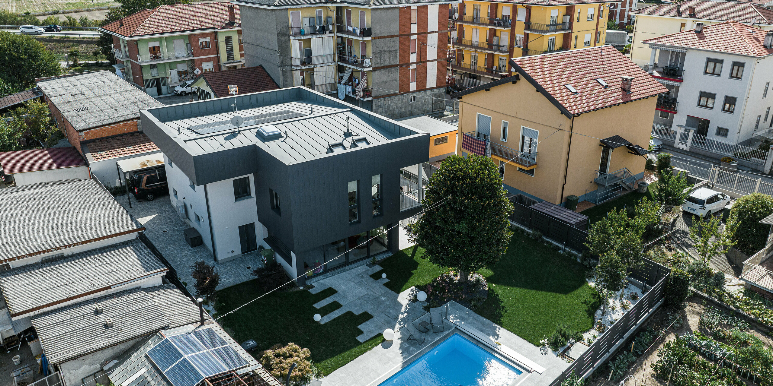 Eindrucksvolle Vogelperspektive eines modernen Einfamilienhauses in Savigliano, Italien, mit einer PREFA Siding.X Aluminiumfassade in P.10 Dunkelgrau. Das Haus zeichnet sich durch ein klares, minimalistisches Design aus und hebt sich mit seiner dunklen Fassade deutlich von der umgebenden traditionellen Bebauung ab. Der großzügige Garten mit einem leuchtend blauen Pool bietet eine attraktive Erholungsoase im städtischen Kontext, während im Hintergrund die Alpen eine malerische Kulisse bilden.